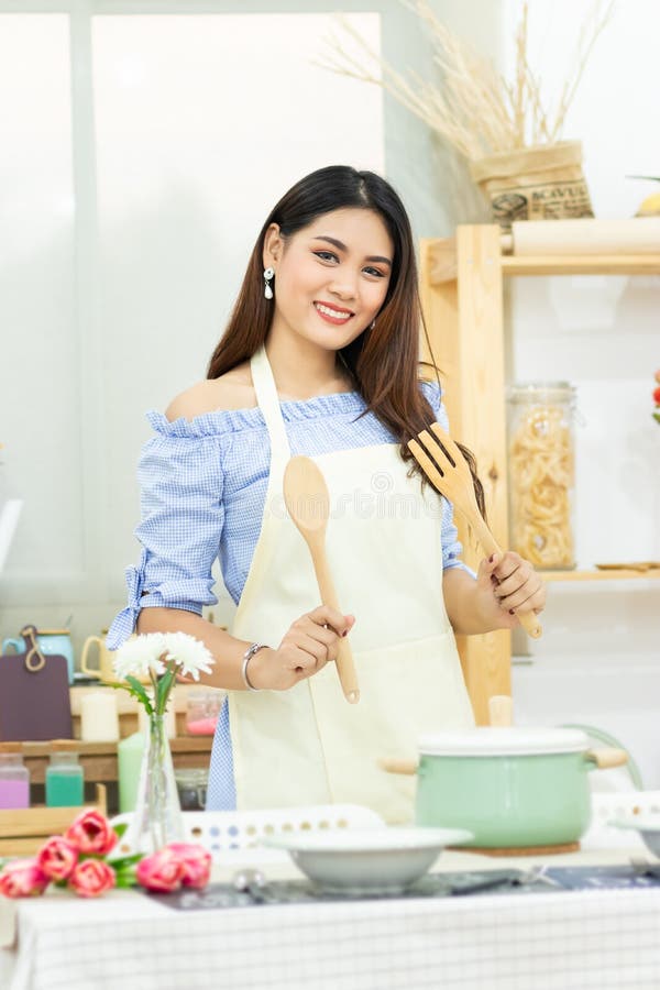 Beautiful Asian woman holding wooden cutlery behind dining table with pot , dish and kitchenware in the kitchen room with smiley stock images