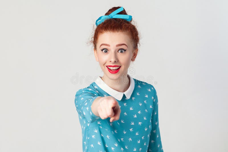 The beautiful redhead girl, wearing blue dress, opening mouths widely, having surprised shocked looks, pointing finger at camera. royalty free stock image