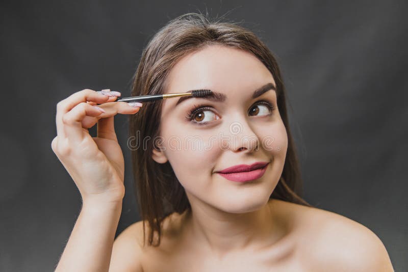 Beautiful young girl stands on a black background. During this, looking away using a brush, makes an eyebrow makeup. Open your mouth stock photo