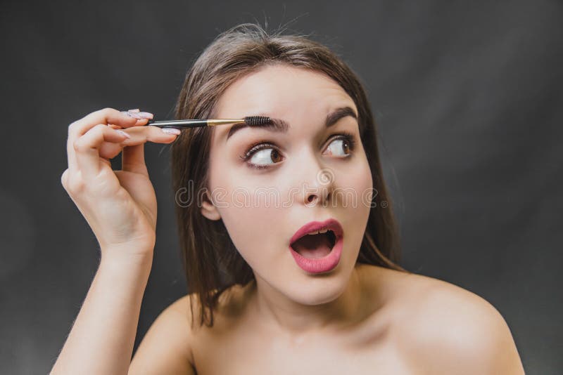 Beautiful young girl stands on a black background. During this, looking away using a brush, makes an eyebrow makeup. Open your mouth stock images