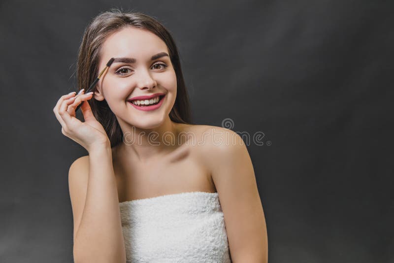 Beautiful young girl stands on a black background. During this, looking into the camera using a brush, makes an eyebrow. Makeup stock photography