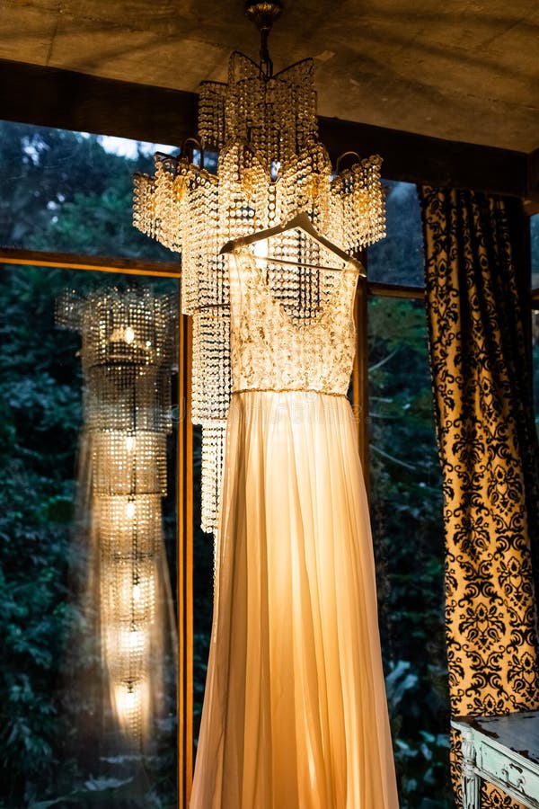 Elegant wedding dress hanging indoors on amazing vintage chandelier. Beautiful warm light stock photography