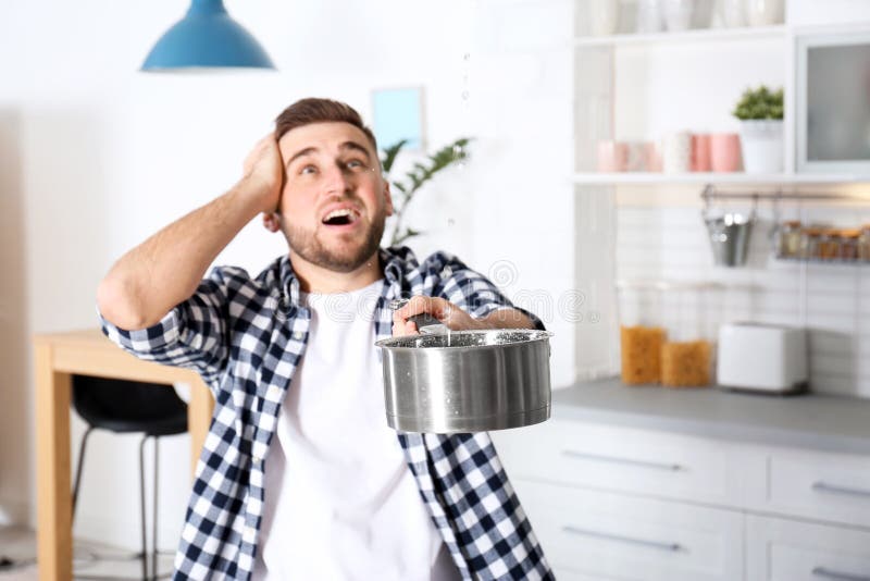 Emotional young man holding saucepan under water leakage from ceiling in kitchen, space for text. Plumber service royalty free stock image