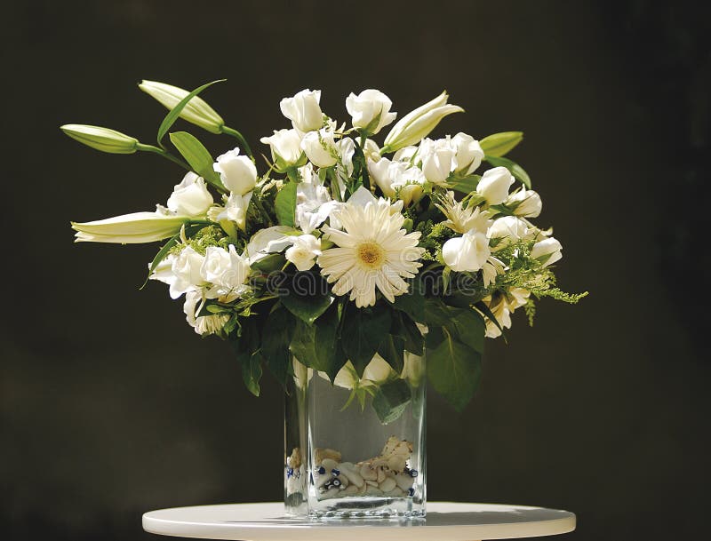 Flowers in Vase. Fresh cut white flowers in clear glass vase, flowers are lilies, roses and daisies, sitting on round small table, dark background stock image