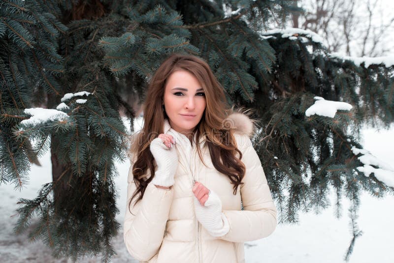 A girl in a winter park on a background of green trees, in a white down jacket with long brown hair, a happy woman. Smiles at coat of lightning royalty free stock photo