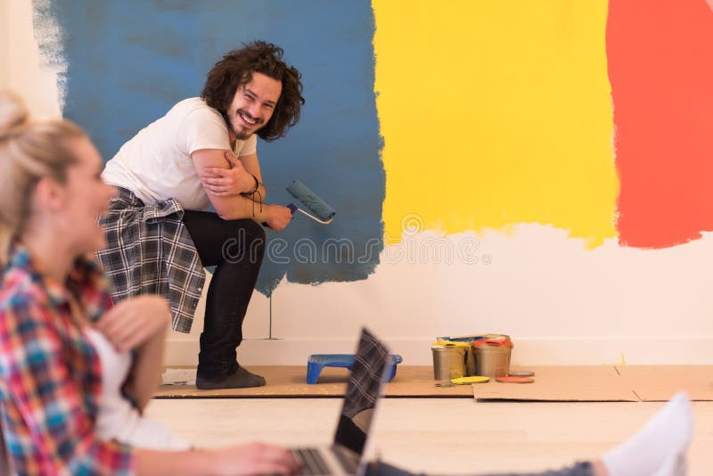 Happy couple doing home renovations. The men is painting the room and the women is relaxing on the floor and connecting with a laptop stock image