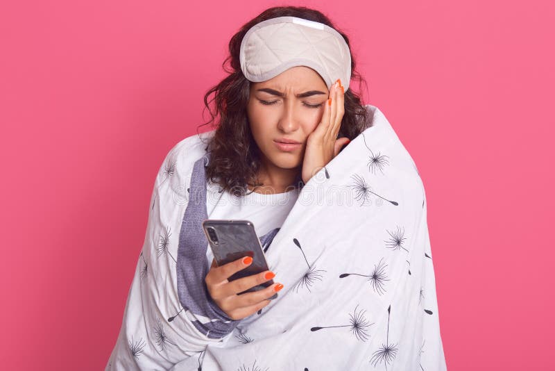 Studio shot of tired exhausted female wrapped in white blanket, having blindfold on her forehead, keeping hand on temple, suffers royalty free stock images