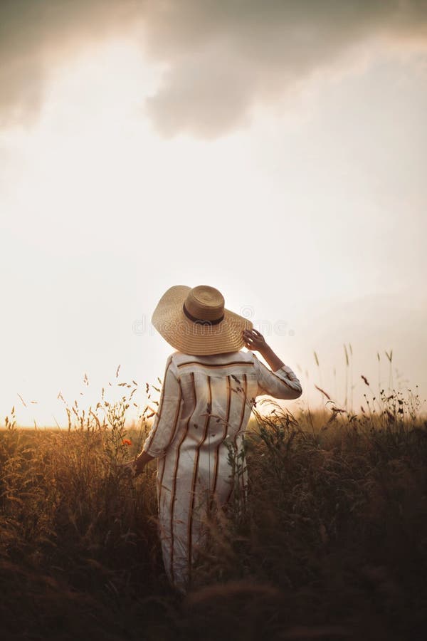 Woman in rustic dress and hat enjoying peaceful sunset in wildflowers and herbs in summer meadow. Atmospheric authentic moment. Stylish girl walking in stock photo