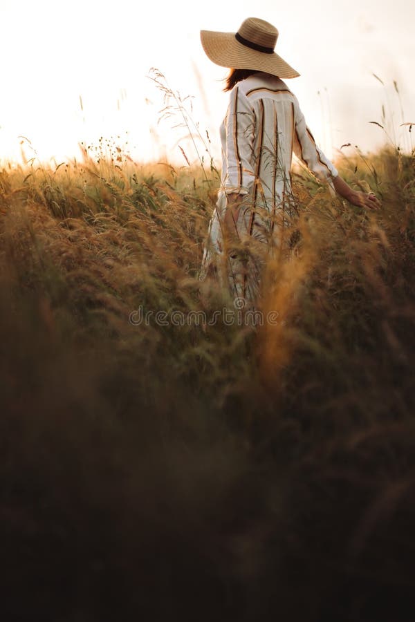 Woman in rustic dress and hat enjoying peaceful sunset in wildflowers and herbs in summer meadow. Atmospheric authentic moment. Stylish girl walking in stock photography