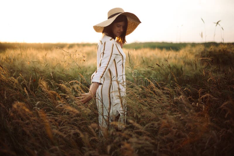 Woman in rustic dress and hat walking in wildflowers and herbs in sunset golden light in summer meadow. Atmospheric authentic. Moment. Stylish girl enjoying stock image