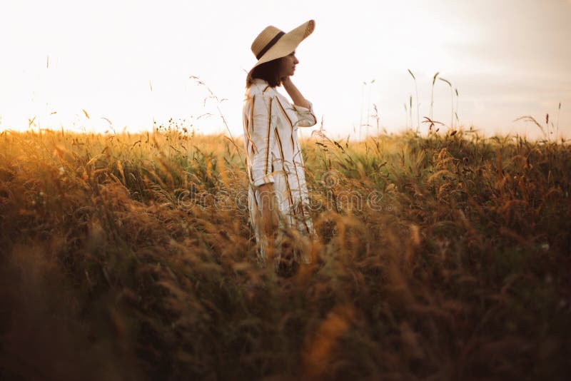 Woman in rustic dress and hat walking in wildflowers and herbs in sunset golden light in summer meadow. Stylish girl enjoying. Evening in countryside. Rural royalty free stock photo