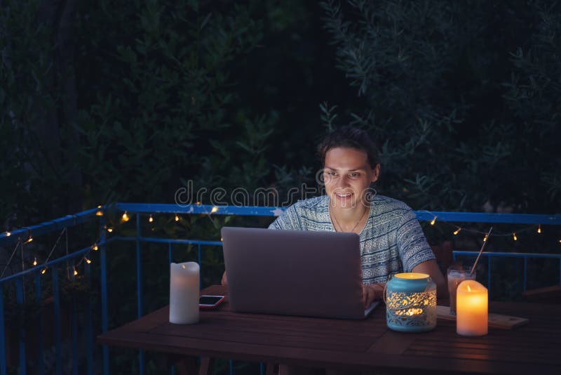 Young hysster woman working on a laptop in the evening on the open terrace of her country house, cozy with candles and lanterns.  royalty free stock photography